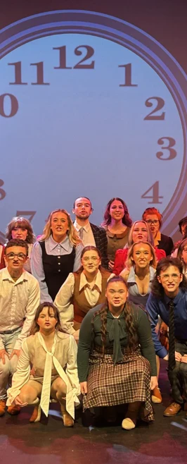 Group of theatre students posing for a cast photo in front of a large projected clock backdrop. Group of theatre students posing for a cast photo in front of a large projected clock backdrop.
